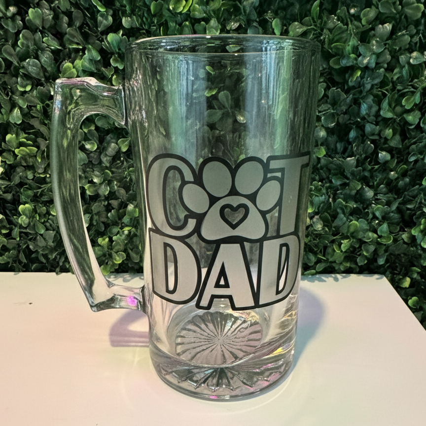 Clear glass beer mug with 'Cat Dad' design etched on surface.  green foliage background
