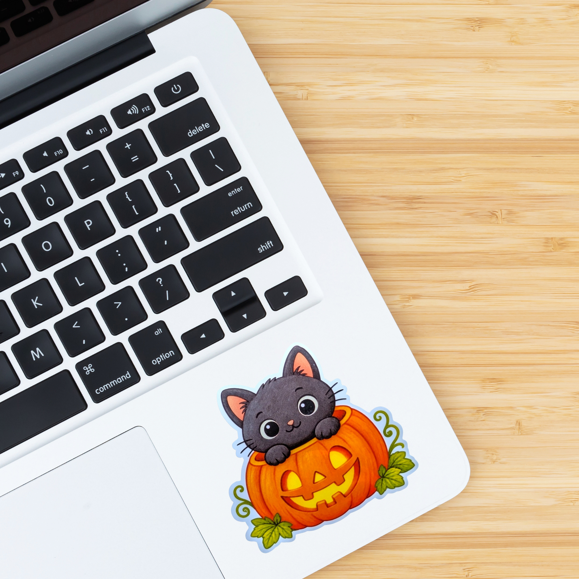 A die-cut vinyl sticker of a cute cartoon cat sitting on a jack-o'-lantern, placed on the palm rest of a white laptop on a light wood grain desk.
