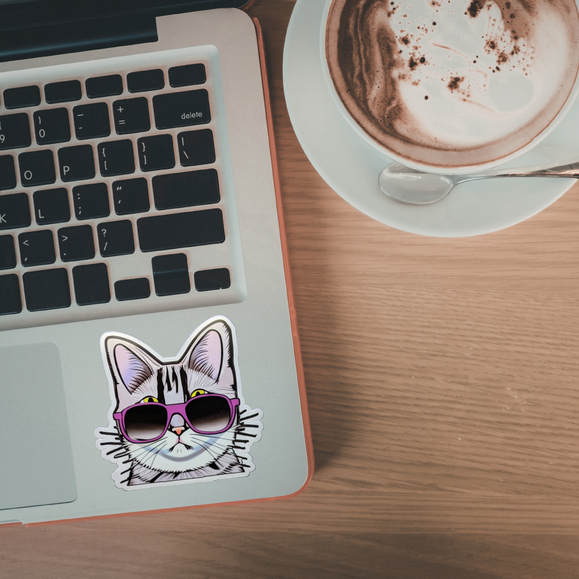 Laptop with a cat sticker wearing sunglasses on a wooden desk next to a cup of coffee.
