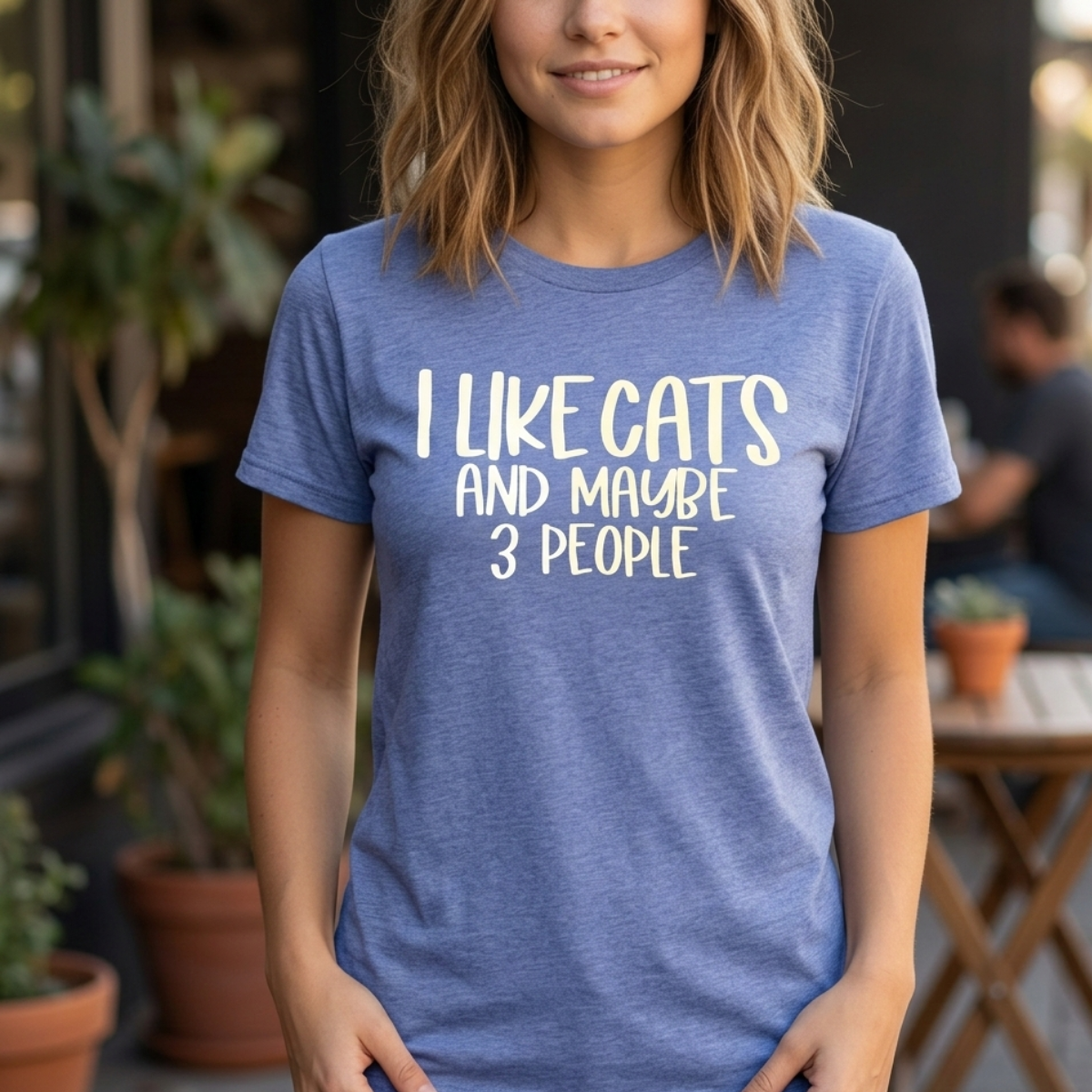 A cropped, medium shot of a woman with medium-length curly hair wearing a blue t-shirt printed with the phrase 'I like cats and maybe 3 people' in white letters. Her face and her waist are cropped.