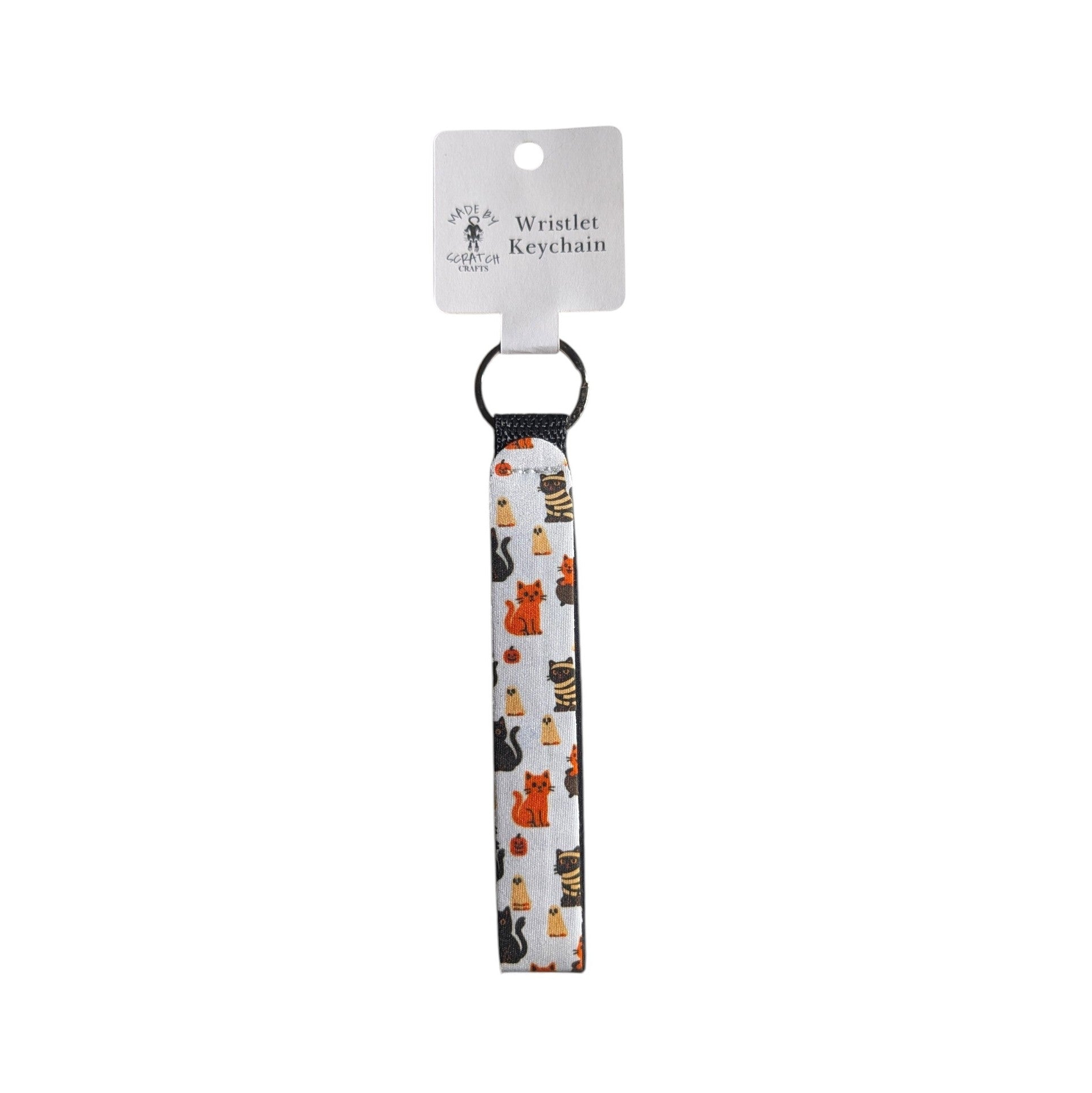 A white wristlet keychain with a black elastic loop and a printed pattern of various cartoon cats in orange, black, and white. It is attached to a white cardboard hanger tag printed with the text 'Wristlet Keychain' and the brand Made By Scratch Crafts.