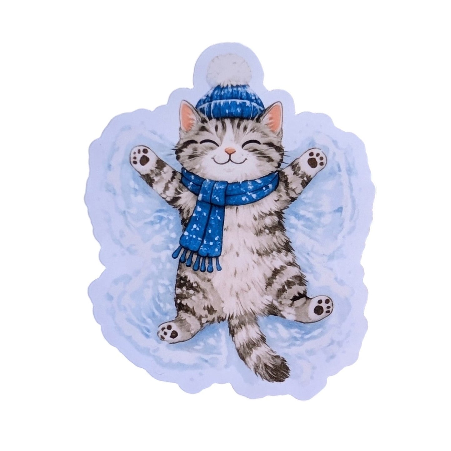 A die-cut sticker or decal featuring an illustration of a gray tabby cat lying on its back in a snow angel pose, wearing a blue knit hat with a pom-pom and a blue starry scarf, set against a light blue snowy background.