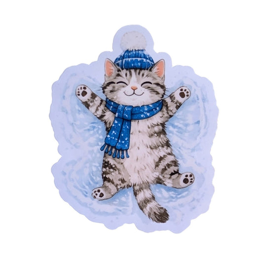 A die-cut sticker or decal featuring an illustration of a gray tabby cat lying on its back in a snow angel pose, wearing a blue knit hat with a pom-pom and a blue starry scarf, set against a light blue snowy background.