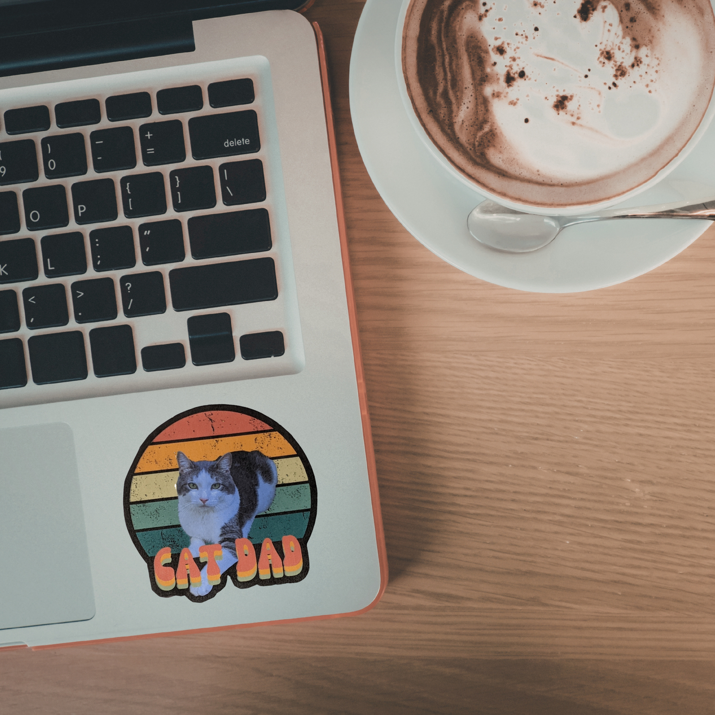 Laptop with a 'Cat Dad' sticker on a wooden desk next to a cup of coffee.