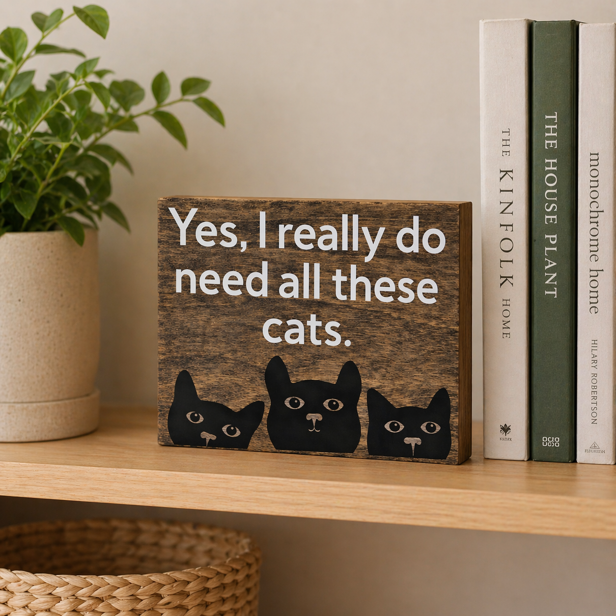 Wooden sign with cat illustrations and text on a shelf with books and a plant.