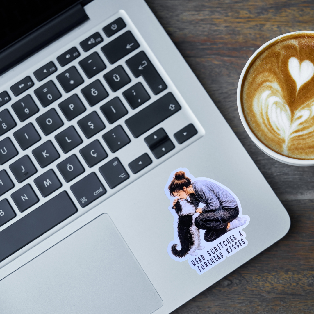 A die-cut sticker featuring an illustration of a woman scratching a black and white cat, placed on the palm rest of a silver laptop next to a cup of latte on a wooden table.