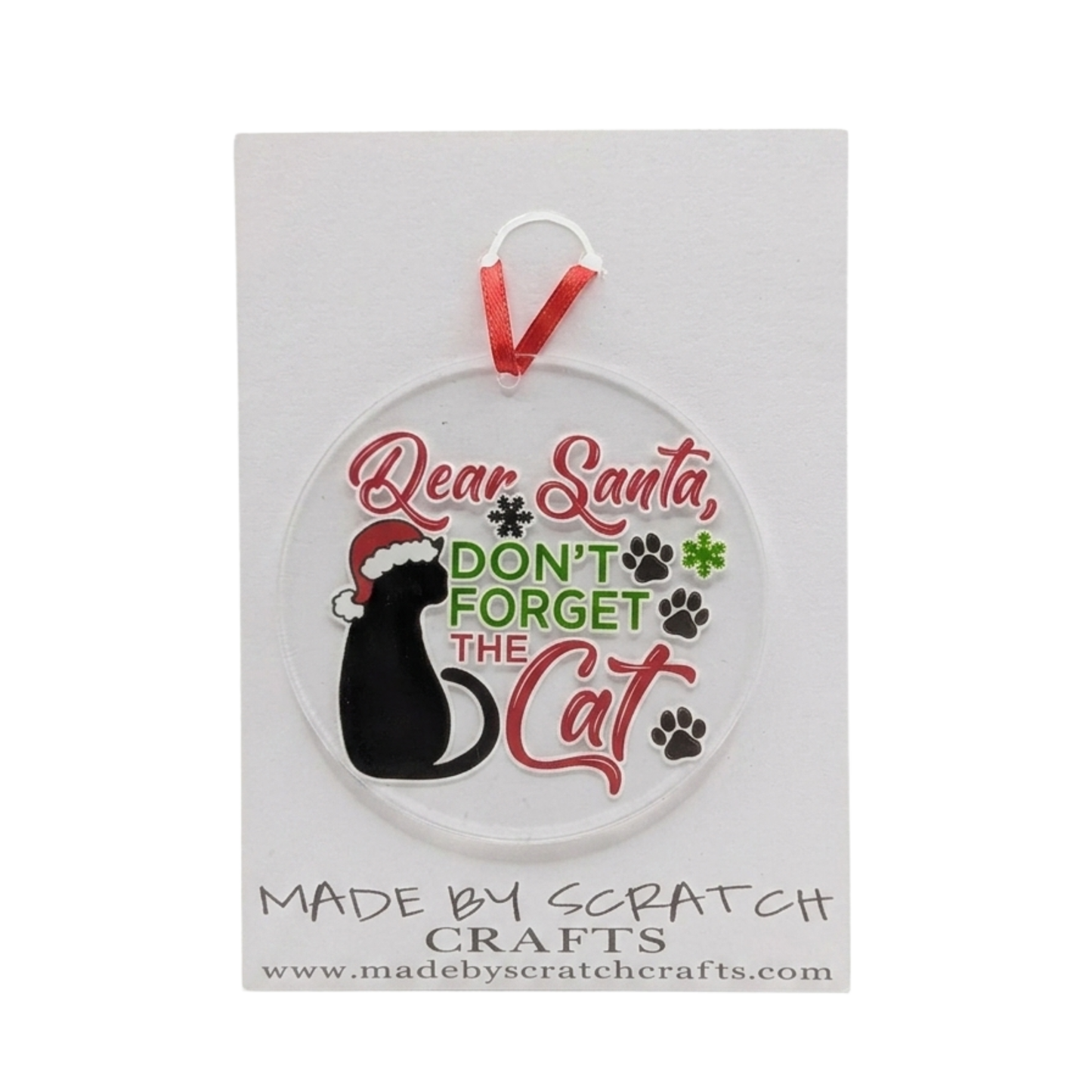A round clear plastic Christmas ornament featuring a black cat wearing a Santa hat, with the text 'Dear Santa, DON'T FORGET THE Cat' in red and green lettering, complete with paw prints. The ornament hangs from a red ribbon and is displayed on a white backing card from Made by Scratch Crafts.