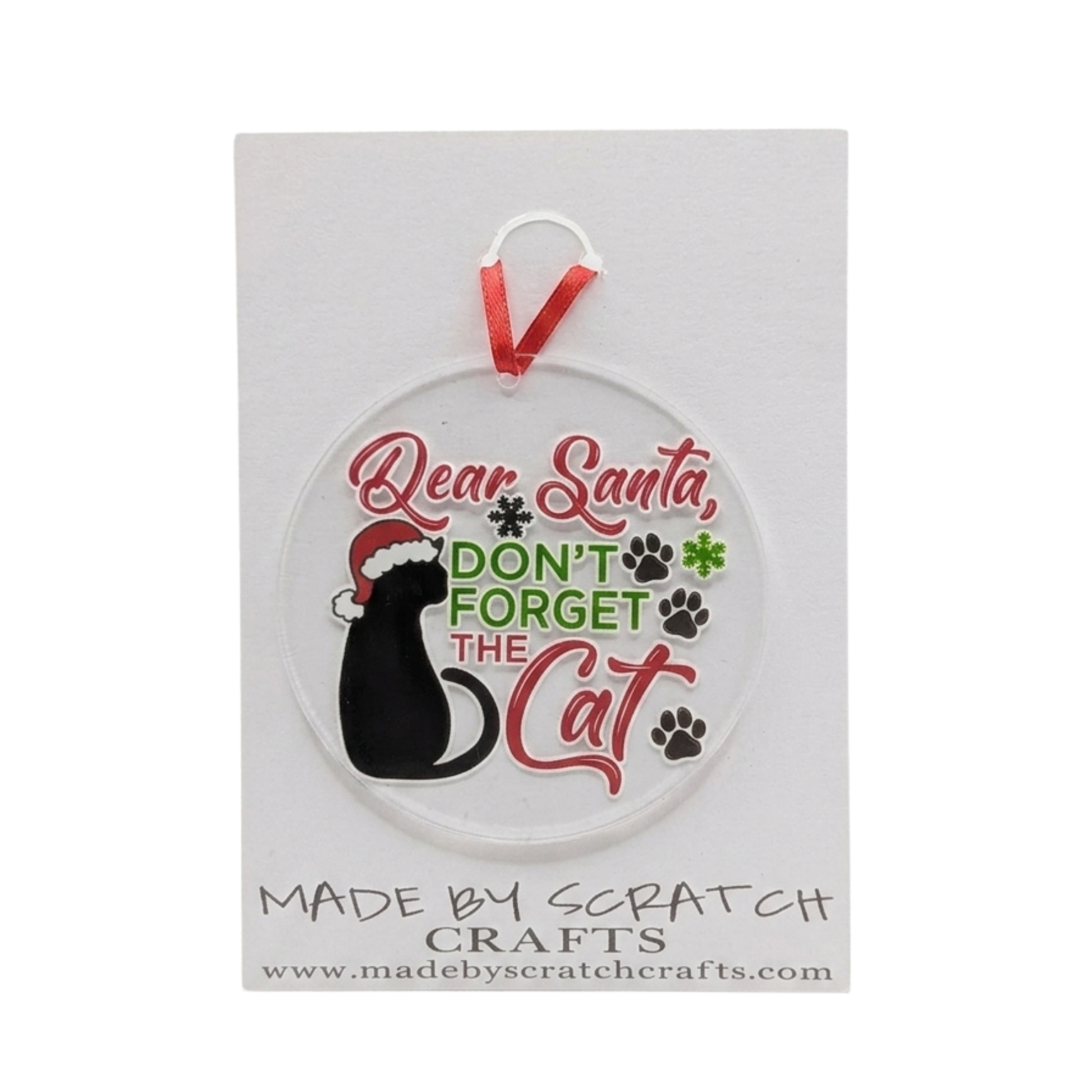 A round clear plastic Christmas ornament featuring a black cat wearing a Santa hat, with the text 'Dear Santa, DON'T FORGET THE Cat' in red and green lettering, complete with paw prints. The ornament hangs from a red ribbon and is displayed on a white backing card from Made by Scratch Crafts.
