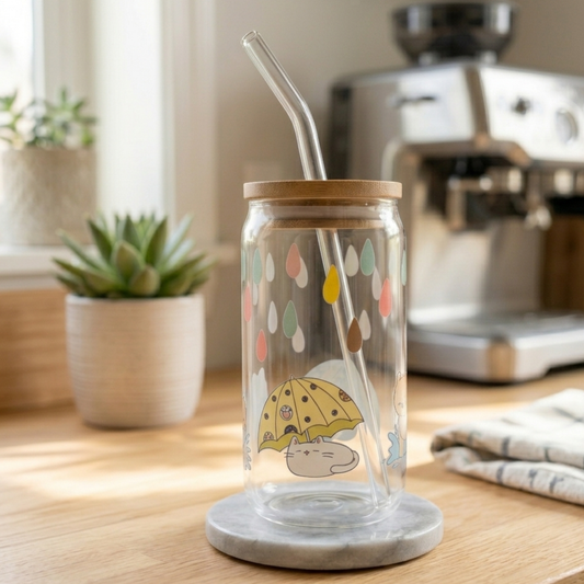 Glass tumbler with straw and raindrop cats design on a marble base, placed on a wooden surface with a coffee machine in the background.