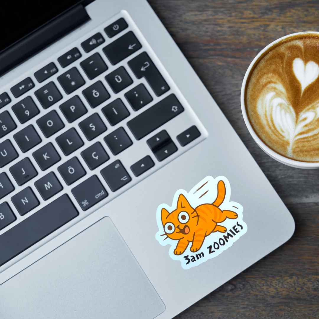 Laptop with a sticker of an orange cat and 'Sam Zoomies' text, next to a cup of coffee on a wooden surface.