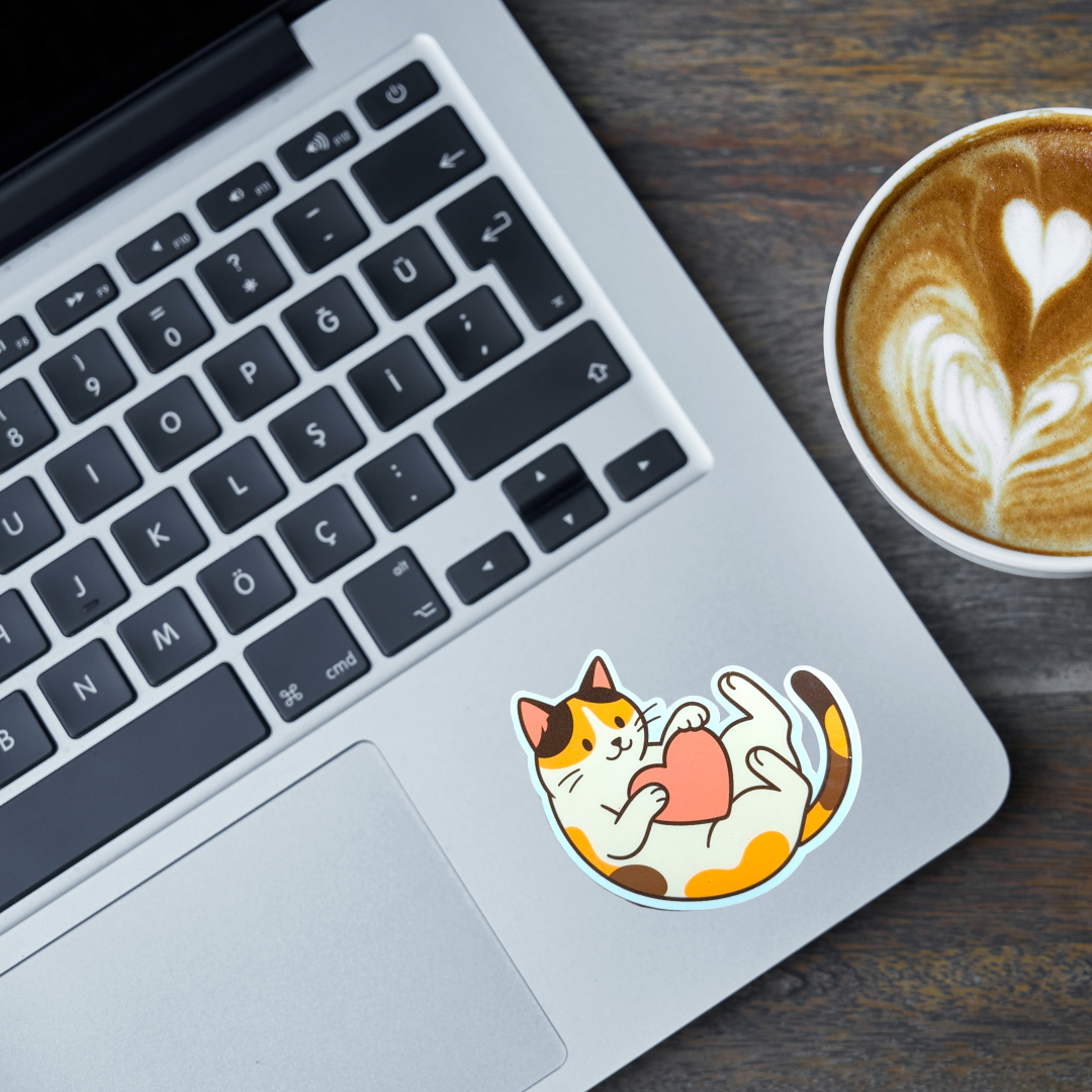 Laptop with a calico cat sticker on the palm rest area next to a cup of coffee.