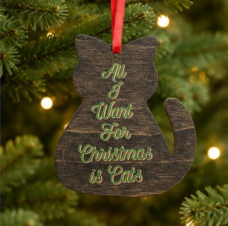 A wooden cat-shaped Christmas ornament hanging on a Christmas tree, with green script text reading 'All I Want For Christmas is Cats' and a red hanging ribbon.