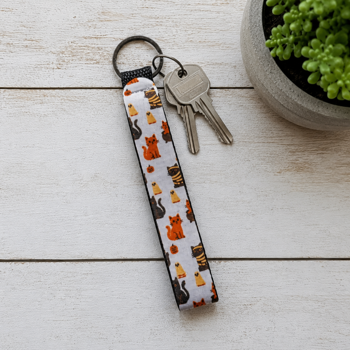 A fabric keychain with a white background and a pattern of orange, black, and yellow cats, attached to a black split ring with two keys. The keychain is laid flat on a white wooden surface next to a small potted green plant.
