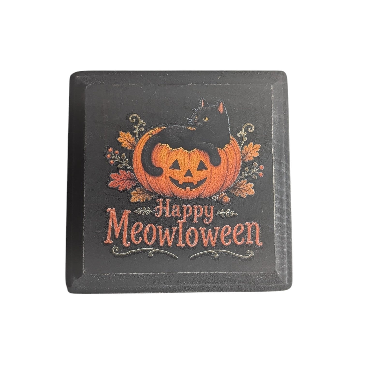 A square black decorative sign featuring a carved orange pumpkin with a black cat sitting on it, surrounded by autumn leaves, and the text 'Happy Meowloween' in orange lettering.