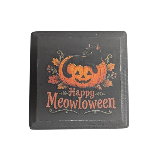 A square black decorative sign featuring a carved orange pumpkin with a black cat sitting on it, surrounded by autumn leaves, and the text 'Happy Meowloween' in orange lettering.