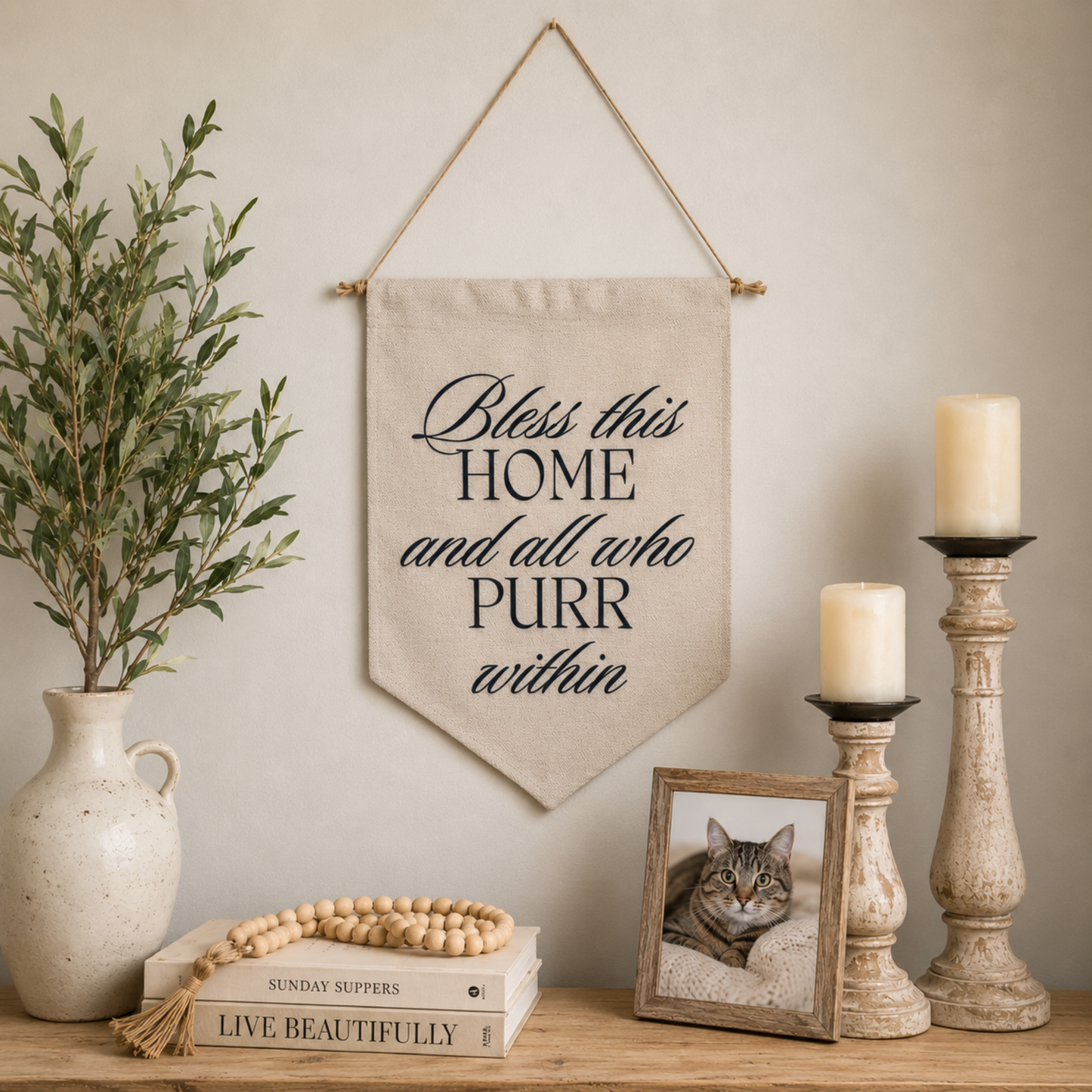 A beige fabric pennant banner hanging on a wall, printed with the phrase 'Bless this HOME and all who PURR within' in dark script. The banner is styled in a cozy home setting on a wooden shelf alongside a potted olive branch in a ceramic vase, a stack of books with a wooden bead runner, a framed picture of a cat, and two distressed candlesticks with lit candles.