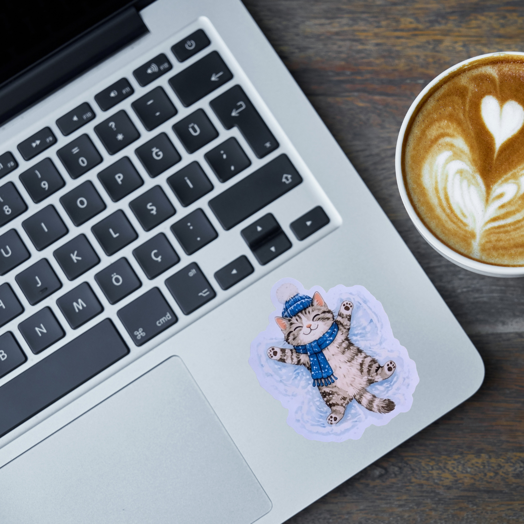 A die-cut sticker of a cat wearing a blue beanie and scarf making a snow angel, lying on a silver laptop palm rest next to a cup of latte art coffee on a dark wooden table.