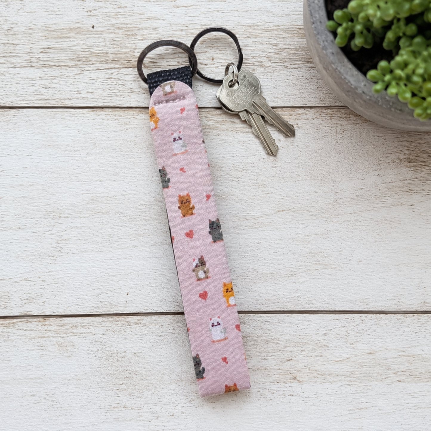 A pink fabric wristlet keychain with a cute cat and heart pattern, holding two keys, displayed on a white wooden surface next to a potted succulent.