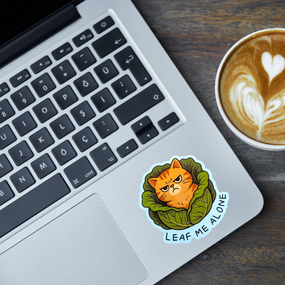 Laptop with a sticker of an angry cat in a cabbage, next to a cup of coffee on a wooden surface.