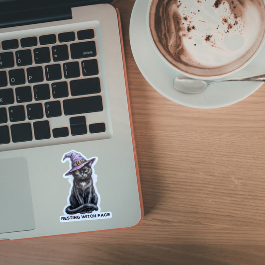 Laptop with a sticker of a cat in a witch hat on a wooden desk next to a cup of coffee.