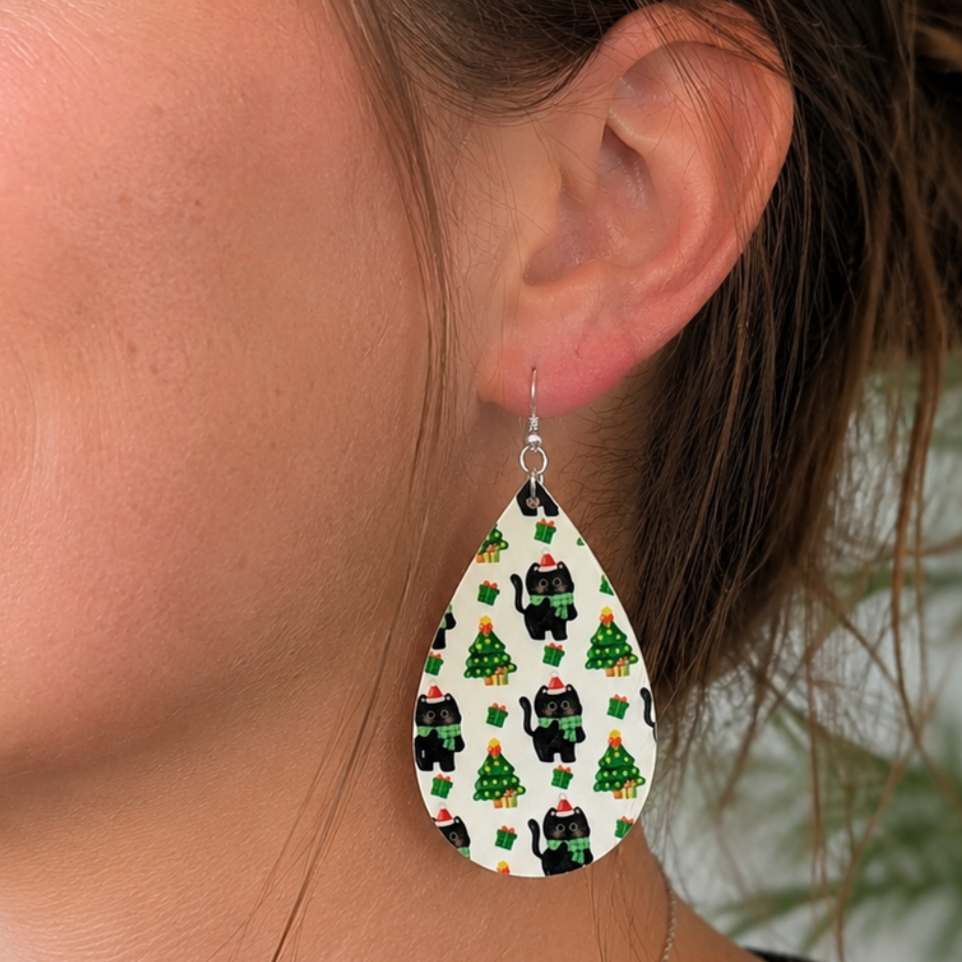 A close-up of a person's ear wearing a large teardrop-shaped earring with a white background and a repeating pattern of black cats wearing green scarves and Santa hats, along with small green Christmas trees.