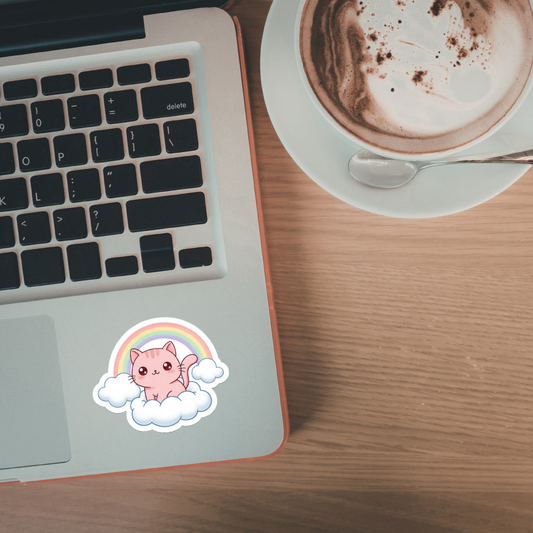Laptop with a cute pink cat sticker sitting on a cloud with a rainbow on a wooden desk next to a cup of coffee.