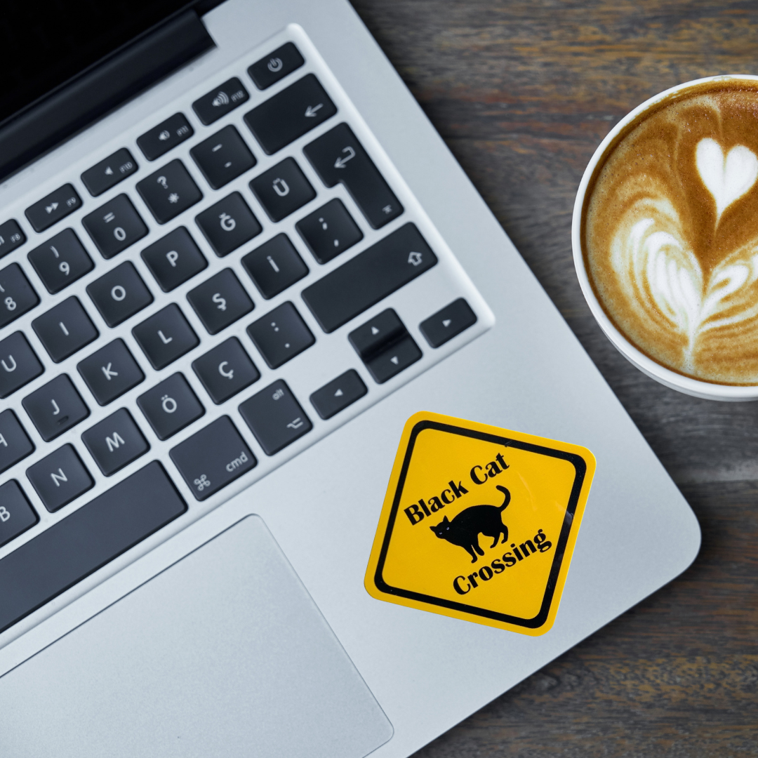 A yellow square sticker with a black border and the words 'Black Cat Crossing' next to a black cat silhouette, shown on a silver laptop next to a cup of latte on a wooden table.
