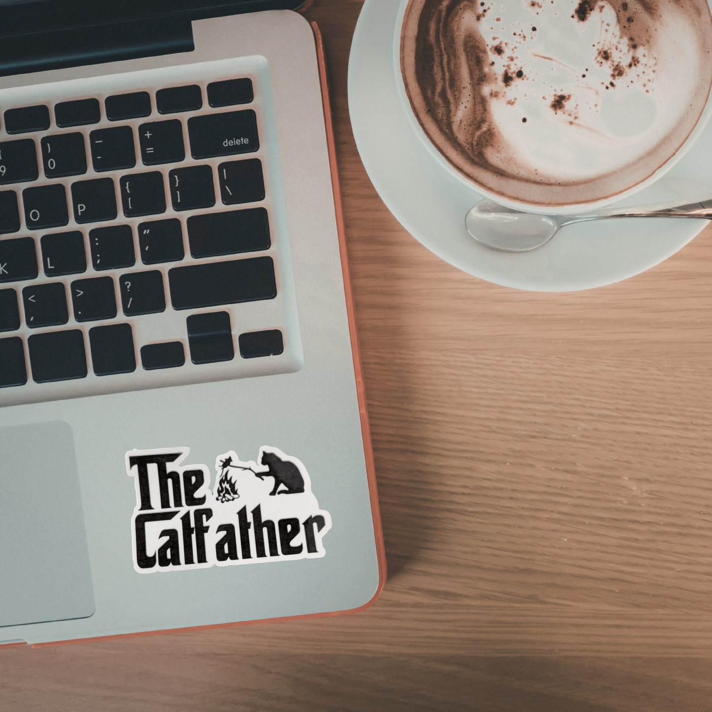 Laptop with 'The Catfather' sticker next to a cup of coffee on a wooden table