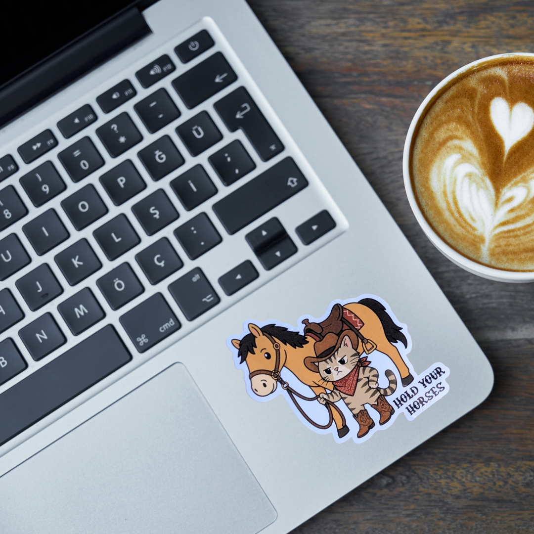 A die-cut vinyl sticker featuring a cartoon cat in a cowboy hat riding a brown horse, with the text 'HOLD YOUR HORSES' printed on the bottom right. The sticker is shown applied to the palm rest of a silver laptop, next to a cup of latte art coffee on a dark wood desk.