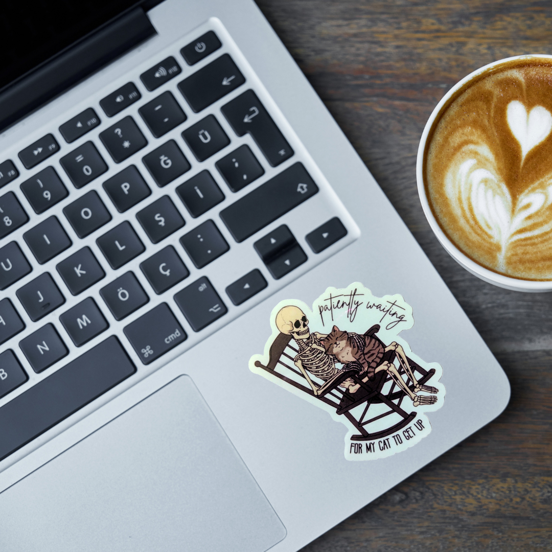 Laptop with a skeleton holding a cat sticker on the keyboard and a cup of coffee with latte art on a wooden surface