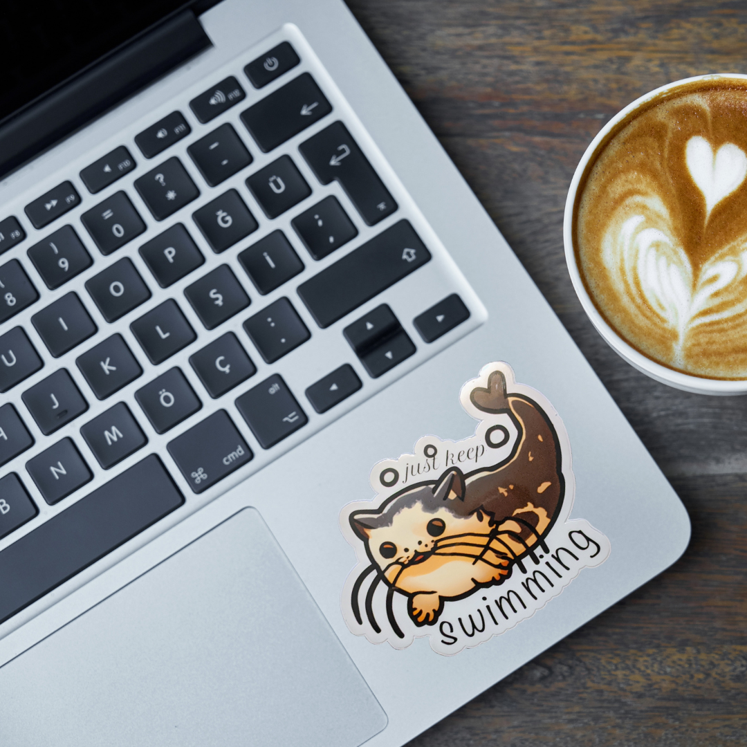 Laptop with a sticker of a catfish and text 'Just keep swimming' next to a cup of coffee on a wooden surface.