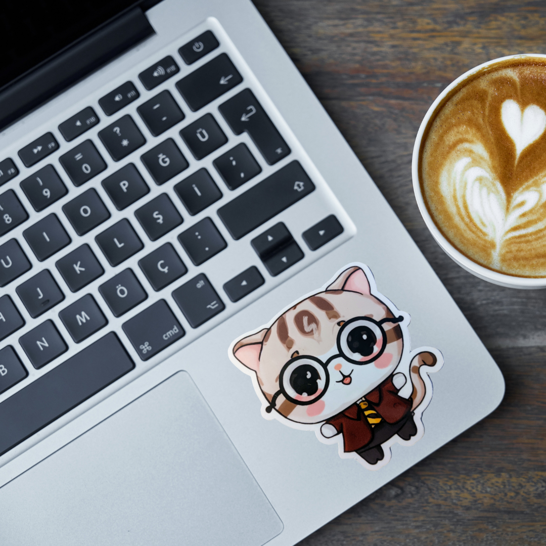 Laptop with a sticker of a cat wearing glasses and a tie, next to a cup of coffee on a wooden surface.