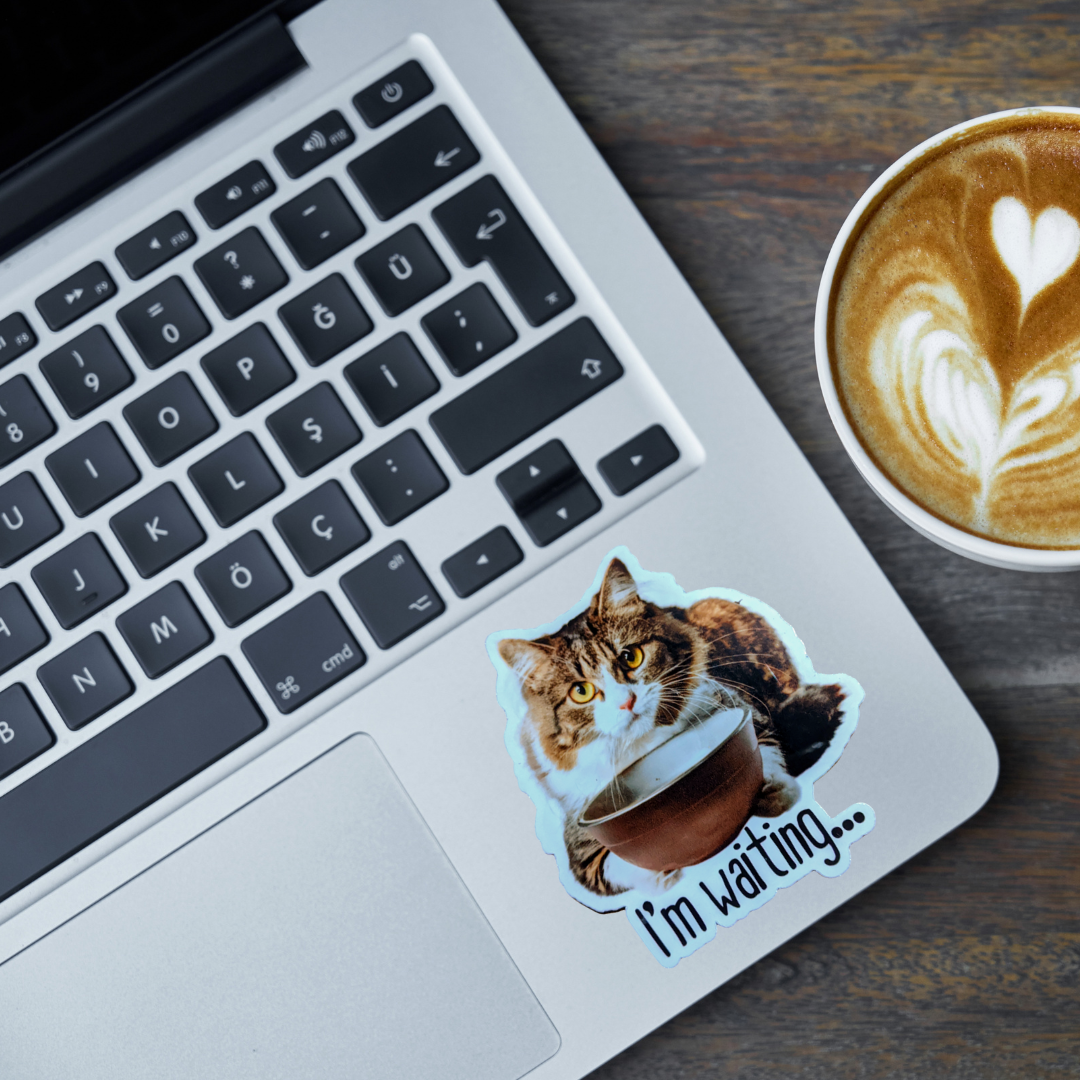 Laptop with a cat sticker saying 'I'm waiting...' next to a cup of coffee on a wooden surface.