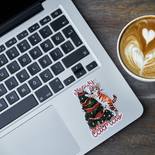 Laptop with a 'Meowy Catmas' cat sticker next to a cup of coffee on a wooden surface