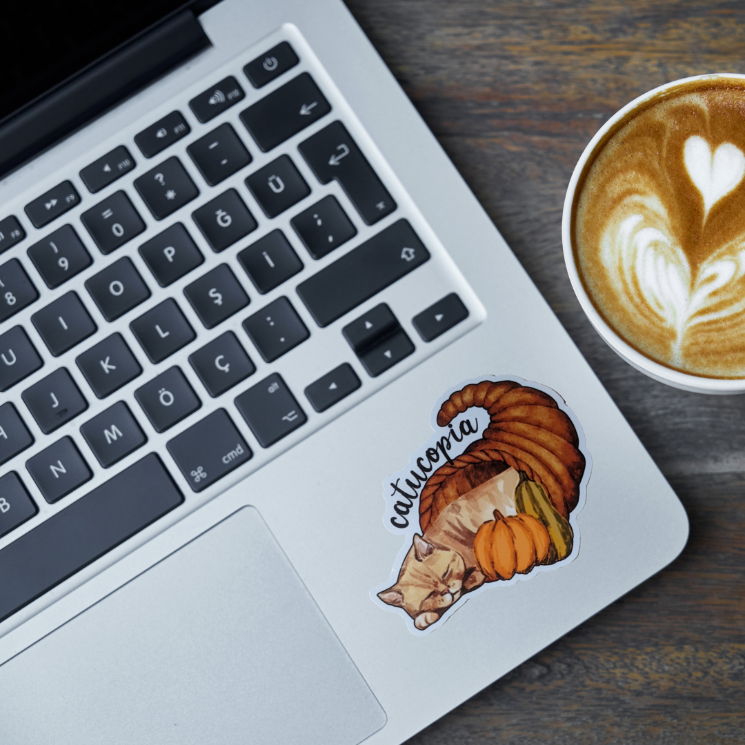 Laptop with a sticker of a cat and cornucopia and pumpkin, next to a cup of coffee on a wooden surface