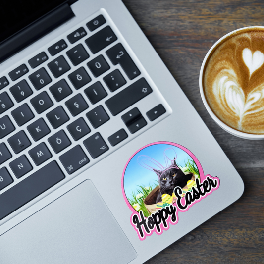 Laptop with a 'Happy Easter' sticker featuring a gray cat wearing rabbit ears, next to a cup of coffee on a wooden surface.