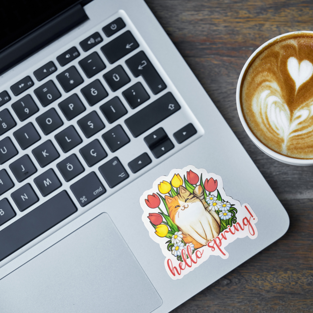 Laptop with a 'hello spring!' cat sticker next to a cup of coffee on a wooden surface