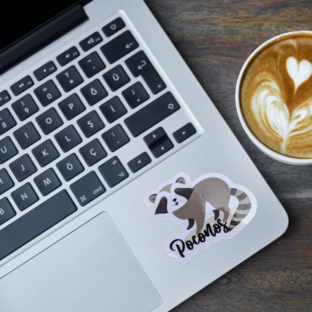 Laptop with a raccoon sticker labeled 'Poconos' next to a cup of coffee on a wooden surface