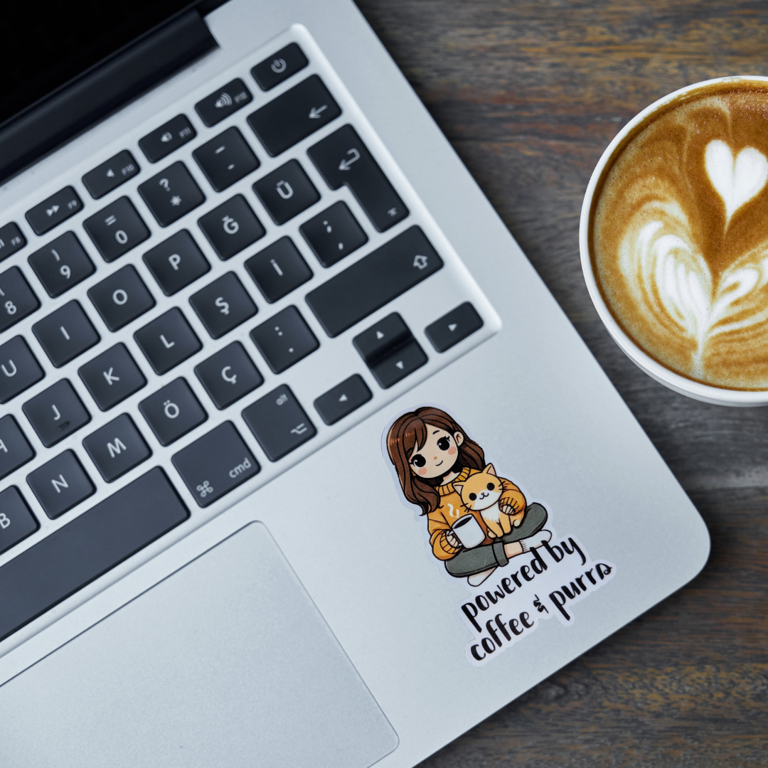 Laptop with a sticker of a person holding a cat and 'powered by coffee & purrs' text, next to a cup of coffee.