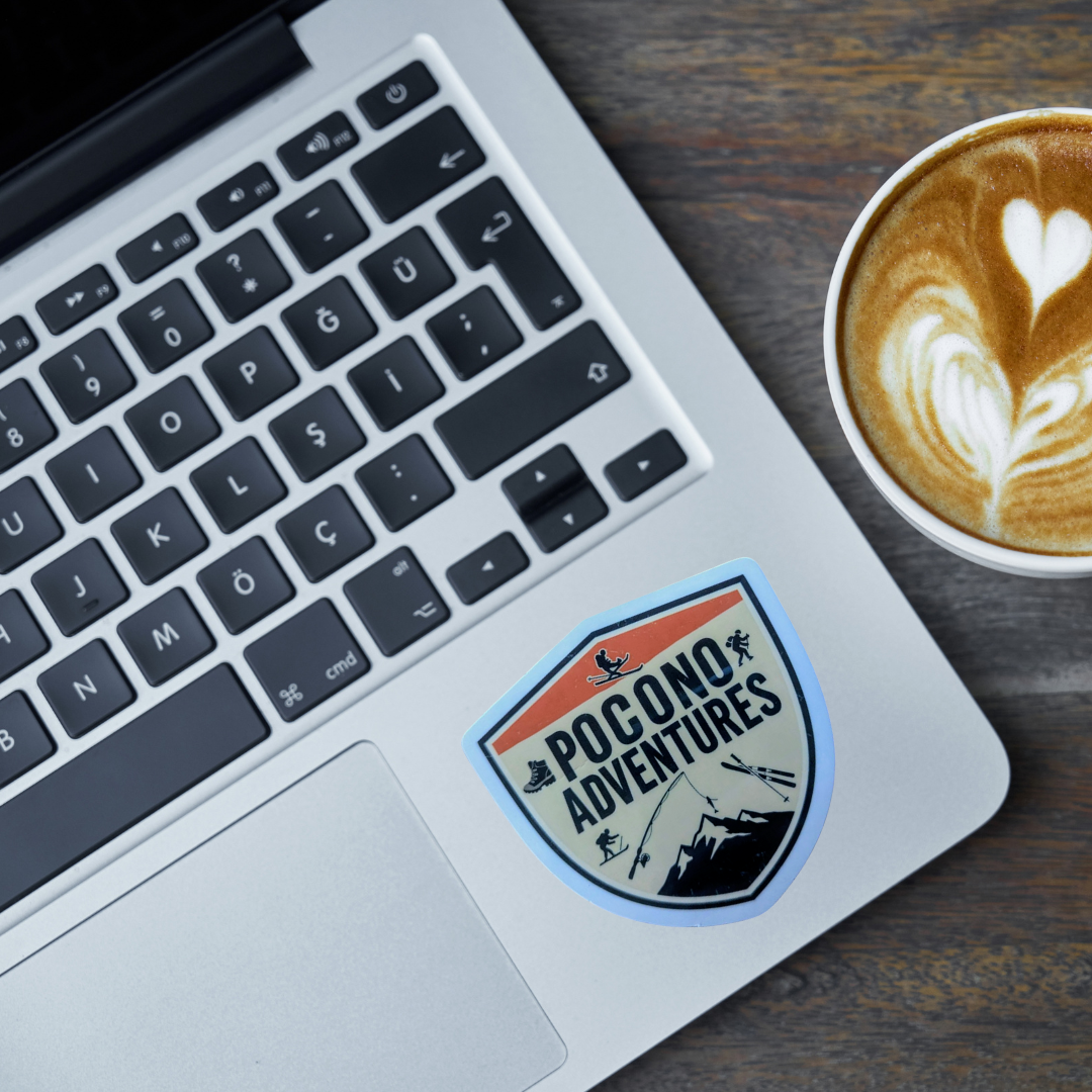 Laptop with a 'Pocono Adventures' sticker next to a cup of coffee on a wooden surface