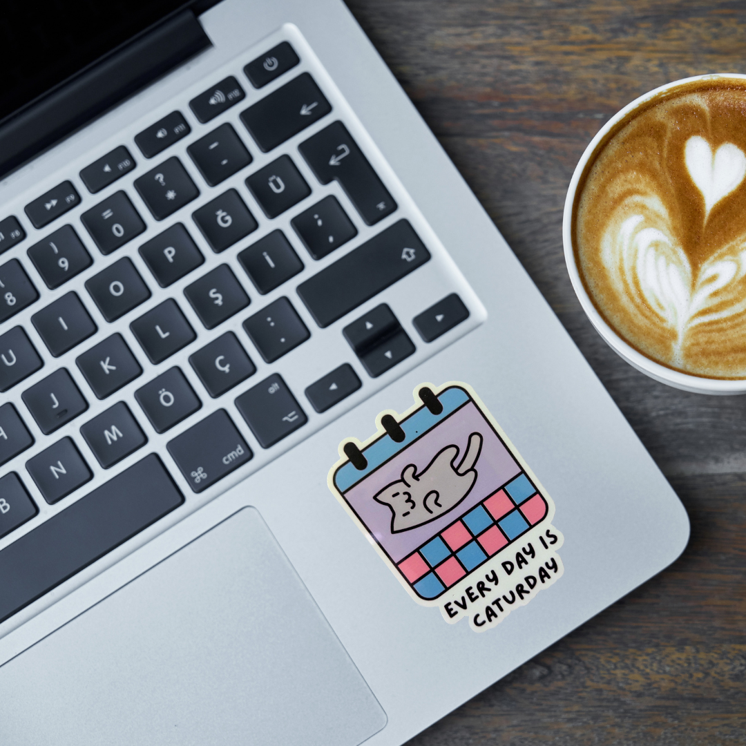 Laptop with a cat calendar sticker on the palm rest area, next to a cup of coffee.