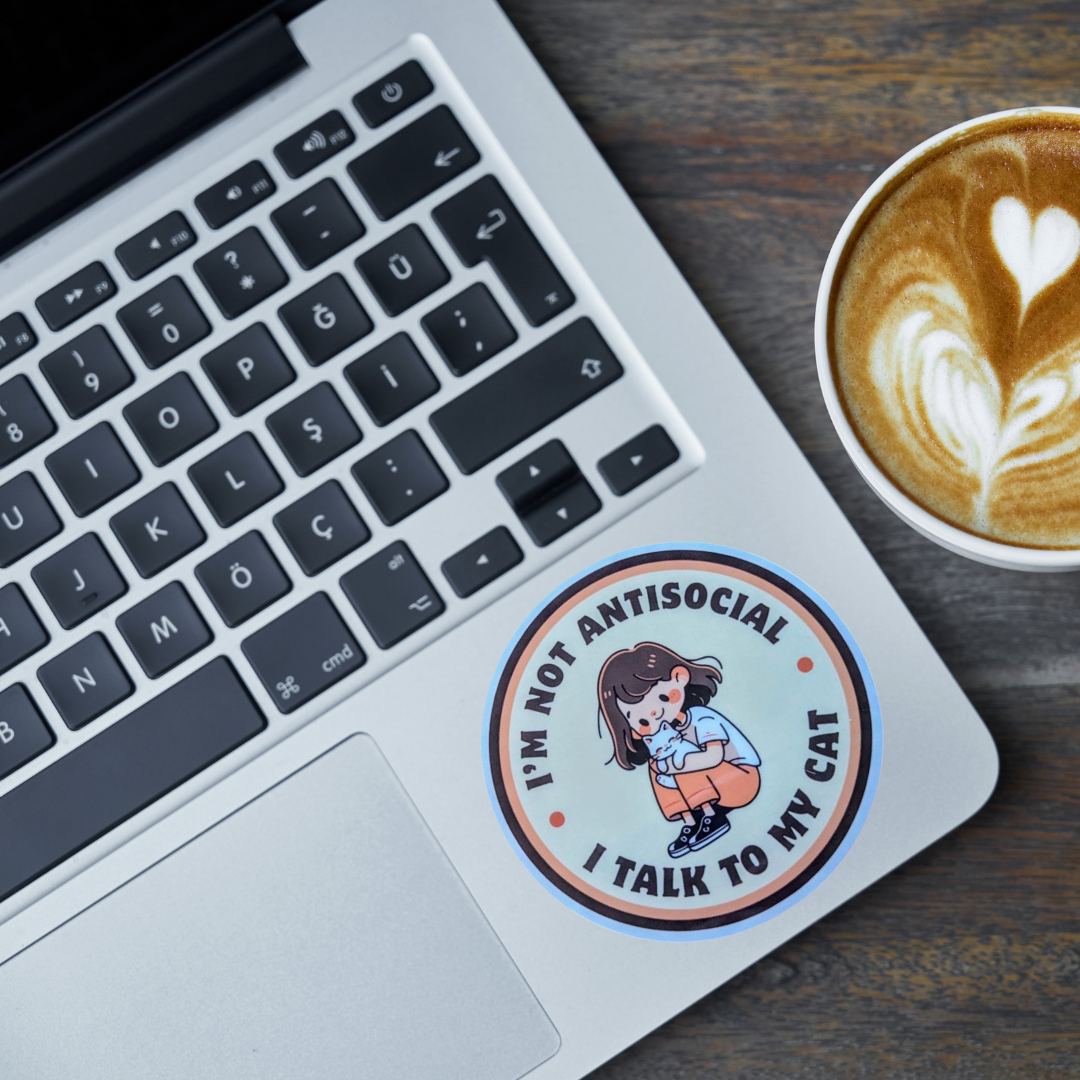 Laptop with a cute sticker of a girl holding a cat that says Im Not Antisocial I Talk To My Cat on a wooden desk next to a cup of coffee
