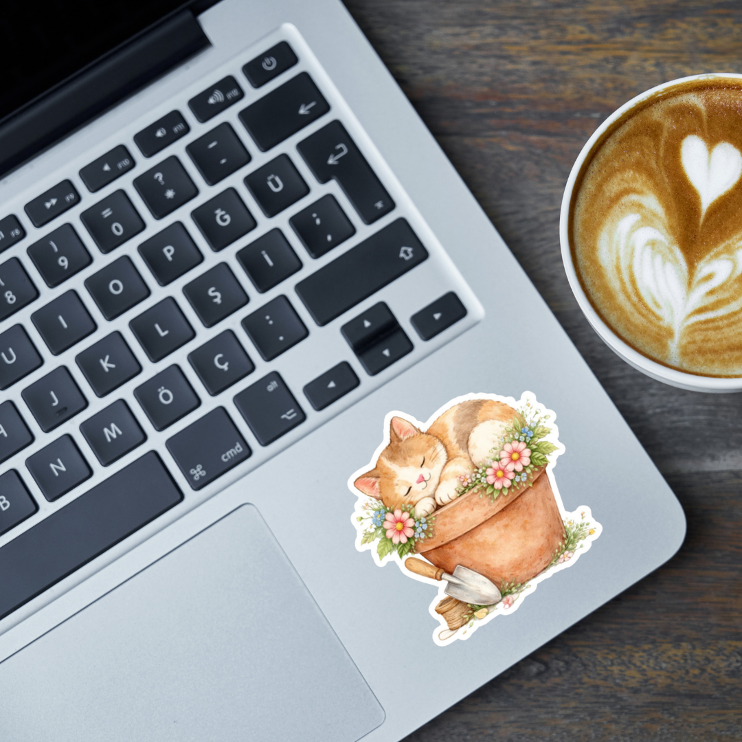 Laptop with a cute cat sleeping in a flower pot sticker on the keyboard next to a cup of coffee.