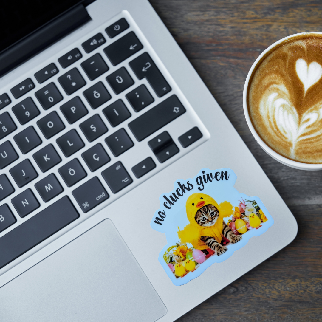 Laptop with a sticker of a cat dressed as a chick and text 'no chucks given' next to a cup of coffee.