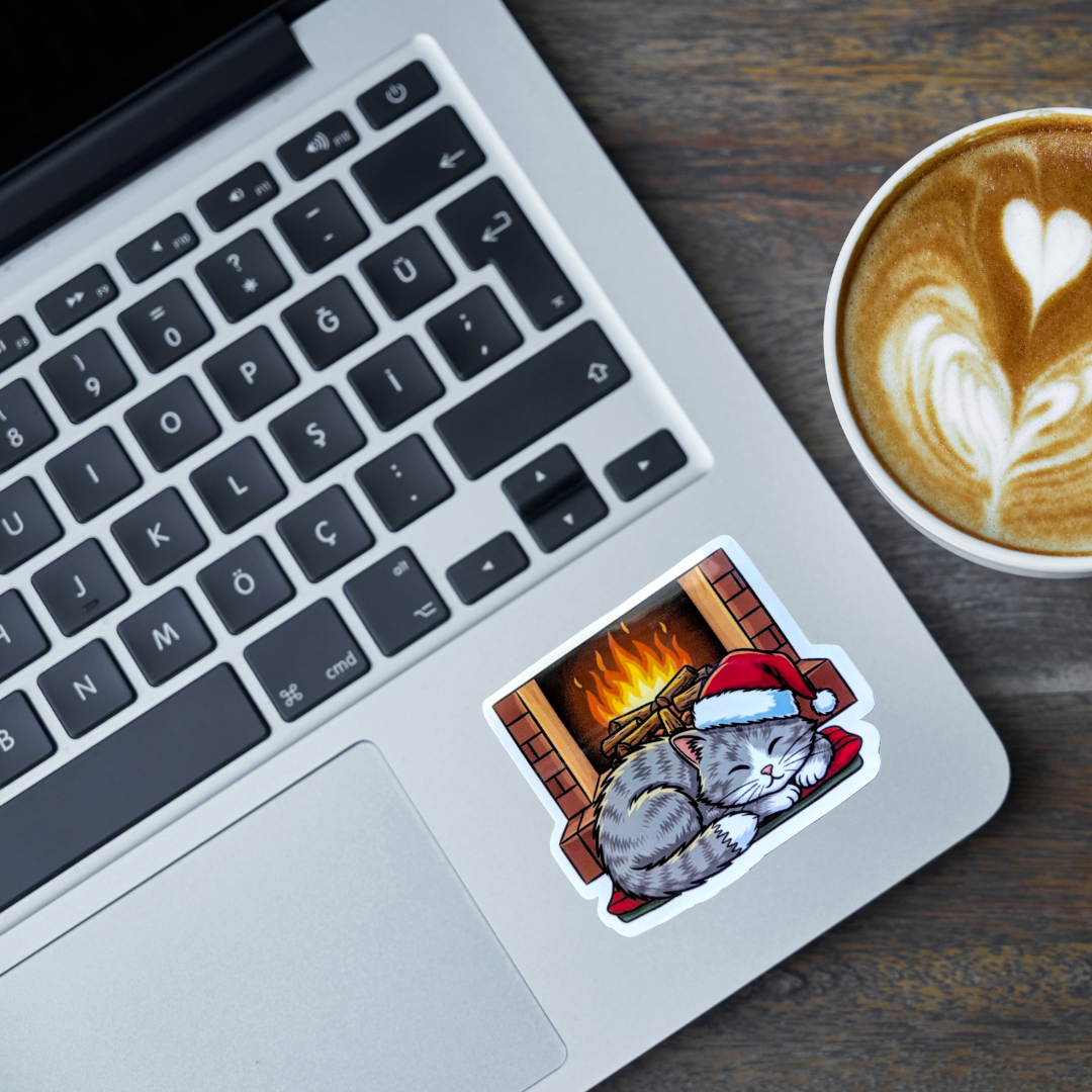 Laptop with a sticker of a cat in a Santa hat by a fireplace, next to a cup of coffee.
